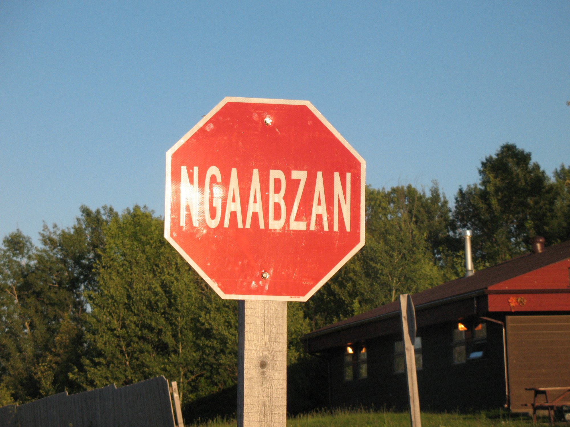 A stop sign in Ojibwe. Its message is one we should all be heeding carefully in these times of accelerational dipshittery. Photo: Hans-Jürgen Hübner, Wikimedia Commons, CC BY 3.0.